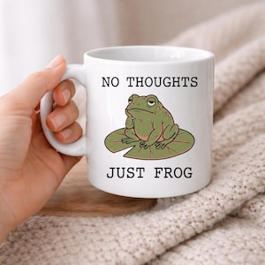 May include: A white ceramic mug featuring a green frog sitting on a lily pad with its hand to its chin. The text above the frog reads NO THOUGHTS and the text below the frog reads JUST FROG in a black sans-serif font. The mug has a handle.