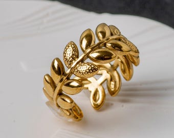 May include: A gold-colored ring featuring a leaf design. The ring has multiple leaves with textured details, creating a nature-inspired aesthetic. The ring is set against a white surface.