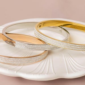 May include: An assortment of elegant bangles in gold and silver tones, adorned with rows of sparkling crystals. The bangles are displayed on a white, shell-shaped plate, creating a sophisticated and glamorous presentation. The jewelry is perfect for adding a touch of sparkle.