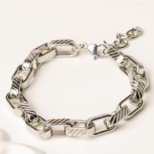 May include: A silver chain bracelet featuring rectangular links with a textured pattern. The bracelet, with a lobster clasp, is shown on a white background. This piece of jewellery is a fashion accessory.