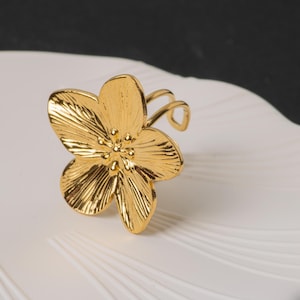 May include: A gold-colored flower-shaped ring. The ring features a detailed floral design with textured petals and a band that wraps around the finger. The ring is set against a white surface with a black background.