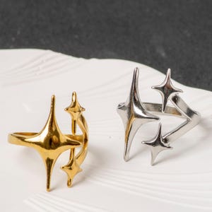 May include: Two rings with starburst designs. One is gold-coloured, the other silver-coloured. They are displayed on a white surface with a dark background. The starbursts have pointed edges.