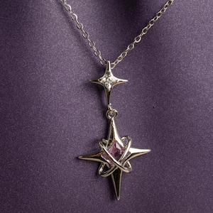 May include: A silver pendant necklace with a starburst design. The necklace has a chain and two star-shaped pendants, one with a small diamond and the other with a pink gemstone. The background is a deep purple.