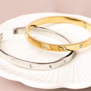 May include: Two hinged bangles, one gold-toned and one silver-toned, rest on a white, scalloped plate. Both bracelets feature small, clear, round gemstones. The gold bangle has a polished finish, while the silver one has a matte appearance.