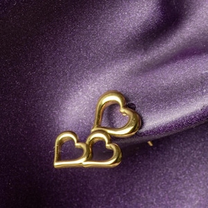 May include: Gold-toned stud earrings featuring a linked heart design. The earrings are set against a textured, deep purple background. The heart shapes are open and have a polished finish.