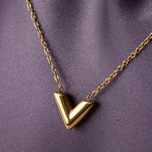 Op de afbeelding: Een goudkleurige ketting met een V-vormige hanger. De hanger is een solide, geometrische vorm en de ketting is delicaat. De ketting wordt gepresenteerd tegen een paarse stoffen achtergrond.