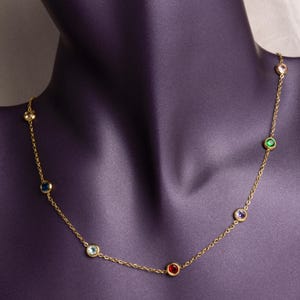 May include: A gold necklace with a fine chain, adorned with a series of colourful gemstones. The necklace features round, bezel-set stones in various colours, including red, blue, green, and pink. It is displayed on a purple mannequin.