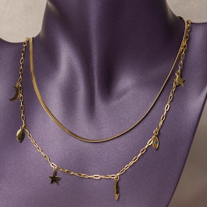 May include: A gold-toned layered necklace featuring two distinct chains. The upper chain is a smooth, flat herringbone style. The lower chain is a delicate oval link chain adorned with small charms including a crescent moon, stars, a lightning bolt, and an eye motif with a blue center.