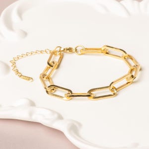 Peut inclure: Un bracelet à maillons de chaîne dorés avec une chaîne et un fermoir réglables. Le bracelet est fait d'un matériau métallique brillant et est présenté sur une surface blanche et décorative. Le bracelet est un bijou.