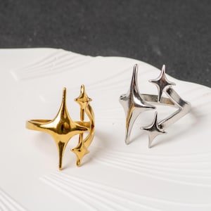May include: Two open-style rings, one gold-toned and one silver-toned, each featuring a multi-pointed star design. The rings have a polished metallic finish and a split band structure, with elongated star shapes extending from the main ring body.