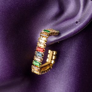 Peut inclure: Boucle d'oreille créole dorée avec une rangée de pierres précieuses rectangulaires colorées. Les pierres sont dans les tons de vert, jaune, rouge, bleu et transparent. La boucle est sur fond violet.