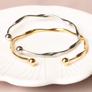 May include: Two open-ended bangles, one silver-toned and one gold-toned, with a twisted design. Each bangle has a small, round ball at each end. The jewelry is displayed on a white, scalloped plate.