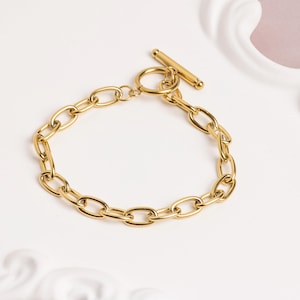 May include: A gold chain bracelet with oval links and a T-bar clasp. The bracelet is presented on a white surface, showcasing its polished finish. The jewellery is designed for everyday wear. The chain links are approximately 1.5 cm in length.