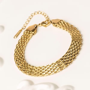 Può includere: Un braccialetto color oro con un design a più fili. Il braccialetto presenta una serie di maglie intrecciate, creando un aspetto strutturato e stratificato. La chiusura e la catena regolabile sono anch'esse dorate. Il braccialetto è esposto su una superficie bianca.