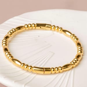 May include: A gold-colored bracelet with a magnetic clasp. The bracelet is made of small, rounded beads and elongated, cylindrical beads. The bracelet is displayed on a white, textured surface.