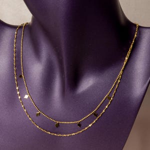 Puede incluir: Un collar doble de oro que presenta dos cadenas distintas. La cadena superior es un delicado hilo de cuentas, mientras que la cadena inferior es de estilo de eslabones trenzados. Ambas cadenas están adornadas con pequeños dijes planos en forma de rombo.