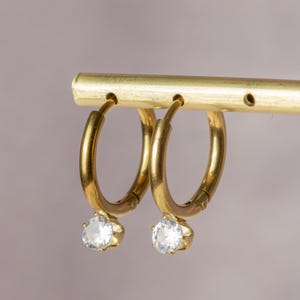 May include: A pair of gold hoop earrings with a clear, round gemstone pendant. The earrings are hanging from a gold bar. The gemstones are faceted and catch the light, creating a sparkling effect.