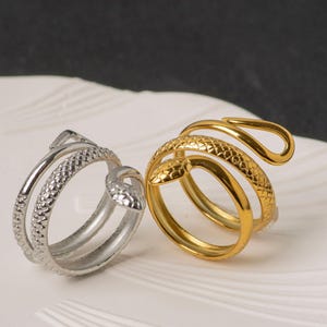May include: Two snake-shaped rings, one silver and one gold, rest on a white surface. The rings feature detailed scales and wrap around the finger. The gold ring has a shiny finish, while the silver ring has a matte appearance.