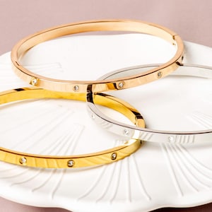 May include: Three gold and silver bangles with small, clear gemstones set into the bands. The bangles are displayed on a white, scalloped plate against a soft pink background. The bangles are different shades of gold and silver.