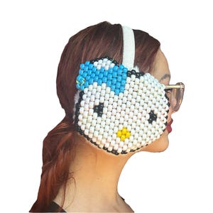 May include: White, black, and blue beaded earmuffs with a cartoon cat face design. The earmuffs have a white headband and a fluffy interior. The cat face features black eyes, a yellow nose, and a blue bow.