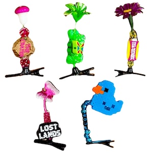 Op de afbeelding: Een verzameling van vijf decoratieve haarclips. Elke clip heeft een uniek ontwerp, waaronder een paddenstoel, een bloem en een gepixelde eend. De clips zijn in verschillende kleuren, met de tekst "LOST LANDS" en "BEATBOX".