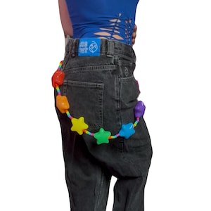 May include: Dark grey denim jeans with a rainbow star-shaped belt. The belt has stars in red, orange, yellow, green, blue, and purple. The jeans have a blue patch on the back pocket with the text "BIG BOY".