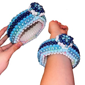 May include: Two beaded bracelets in shades of blue, white, and dark blue. One bracelet is held in a hand, the other is worn on a wrist. The bracelets are made of small, round beads and have a cuff-like design. The bracelet on the wrist has a flower-like embellishment.