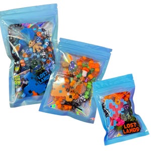 Könnte beinhalten: Drei transparente Zip-Lock-Beutel, gefüllt mit farbenfrohem Perlschmuck und Anhängern. Die Beutel enthalten Armbänder mit verschiedenen Perlen, darunter Buchstaben, pixelige Designs und kleine Figuren. Der Text "LOST LANDS" ist auf zwei der Beutel zu sehen.