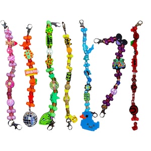 Puede incluir: Una colección de coloridos llaveros de cuentas con varios dijes. Los llaveros presentan una variedad de cuentas en rosa, naranja, verde, amarillo, azul y morado. Los dijes incluyen una bola de discoteca, dinosaurios y un pato azul.