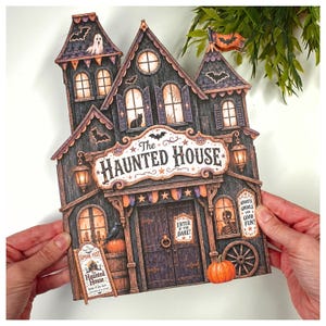 May include: A flat, die-cut paper decoration shaped like a haunted house with dark wood textures, orange pumpkins, bats, and a ghost. The sign reads The Haunted House, Enter If You Dare, and Ghosts Ghouls and Good Fun in a detailed, illustrated style.