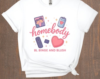 May include: White t-shirt with a graphic design featuring the word "homebody" in pink script, along with illustrations of a photo, a can with a heart, a remote, a memory card, and a pink heart. The text "BL BINGE AND BLUSH" is below.