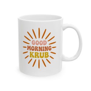 May include: White ceramic mug with the text "GOOD MORNING KRUB" in pink, orange, and yellow, surrounded by orange sun rays. The mug has a curved handle and is suitable for hot or cold beverages.