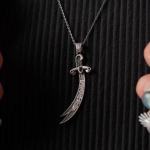 May include: A silver-toned pendant shaped like a curved sword or scimitar, featuring intricate engraved patterns and small black stone accents on the hilt and crossguard. The pendant hangs from a delicate silver-toned chain link necklace.