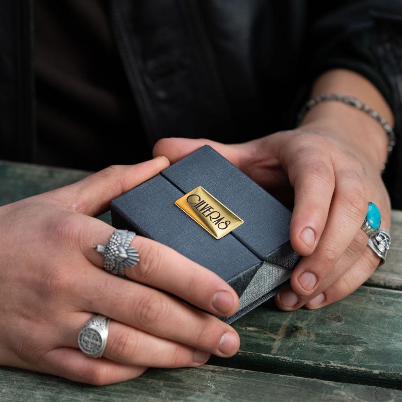 A person's hands holding a small black box with a gold label, wearing multiple rings on their fingers.