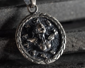 May include: A circular silver-toned pendant featuring a high-relief sculpture of a figure with an open mouth, surrounded by multiple small skulls. The pendant has a textured, darkened background and a decorative braided border, suspended from a metal loop.