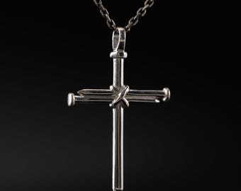 May include: A silver-toned metal cross pendant suspended from a matching chain. The cross is constructed from two intersecting nail-shaped pieces, with a small, twisted metal wire detail wrapped around the center where the two pieces meet.