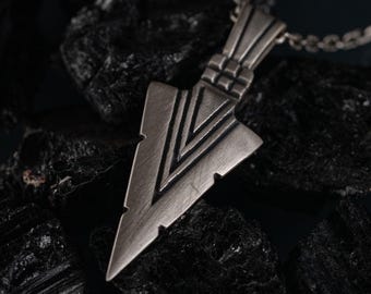 May include: A silver-toned metal pendant shaped like an arrowhead with a brushed finish and engraved V-shaped lines. The pendant features a textured bail at the top and is attached to a matching silver-toned metal chain link necklace.