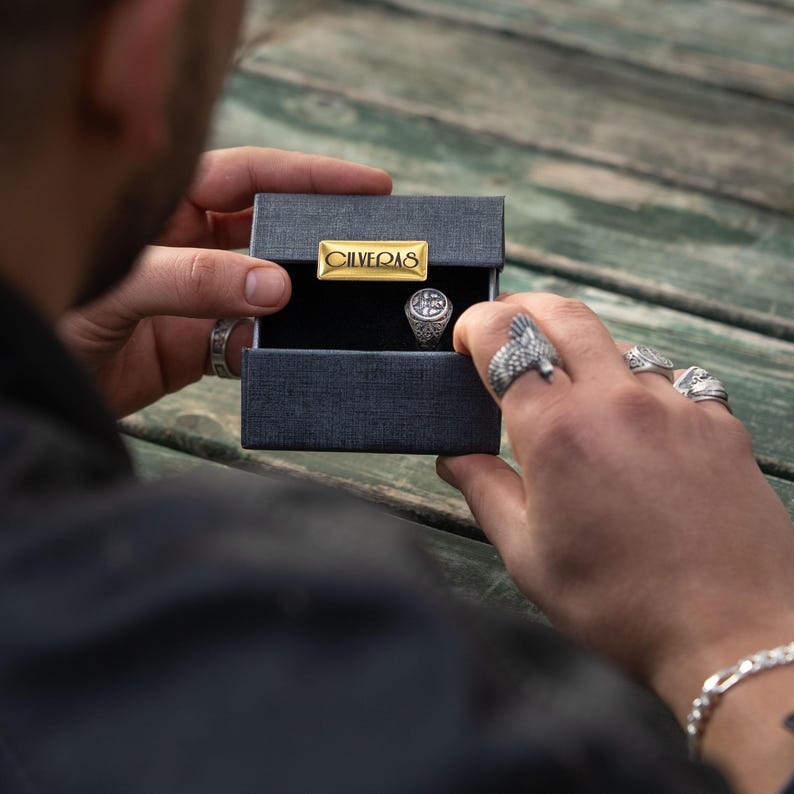 A person is holding a black box with a gold-colored label that says "CLIVEQS" on it, and they are wearing multiple rings on their fingers.