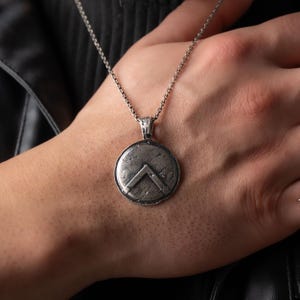 May include: A circular silver-toned pendant featuring a raised inverted V-shaped emblem on a textured, weathered surface. The pendant hangs from a thin, matching silver-toned metal chain, showcasing a detailed, oxidized finish on the metal.