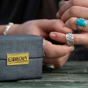 A person's hands wearing multiple rings, including a turquoise ring, are shown in close-up, with a box labeled "CILVERAS" in the foreground.