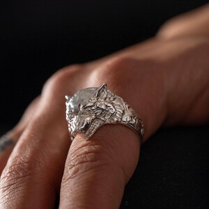 May include: A silver-toned ring featuring a detailed, three-dimensional wolf head design. The metal surface displays intricate fur textures and facial features, including pointed ears and a snout, extending into a band with embossed decorative patterns.
