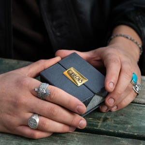 A person's hands holding a small black box with a gold label, wearing multiple rings on their fingers.