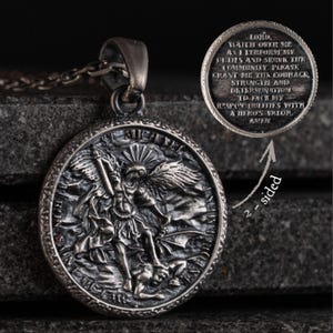 May include: A silver-toned, two-sided pendant necklace. One side depicts an angel slaying a demon, the other side features an inscription. The pendant is round with intricate detailing and hangs from a delicate chain. The text on the pendant reads "2-sided".