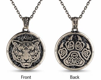 May include: A silver-toned pendant necklace featuring a circular design. The front displays a detailed tiger face, while the back shows a stylized animal paw print with intricate knotwork patterns. The metal has an oxidized finish and a chain.