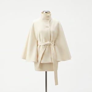 Peut inclure: Manteau cape crème avec un col montant et une ceinture assortie. Le manteau est doté d'une fermeture boutonnée et de manches larges. La ceinture est nouée à la taille, et le manteau est présenté sur un mannequin sur fond blanc.