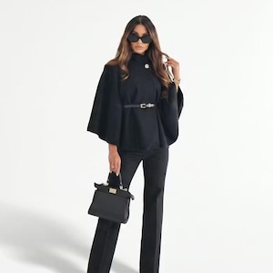 Peut inclure: Un haut noir de style cape avec un col montant, associé à un pantalon noir assorti et une ceinture noire. La tenue est accessoirisée d'un sac à main noir et de lunettes de soleil. L'ensemble est présenté sur un fond blanc, créant un look monochrome élégant.