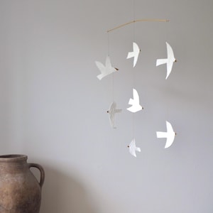 Op de afbeelding: Een handgemaakte mobiel met witte papieren vogelvormen die aan een houten stok hangen. De vogels hangen op verschillende niveaus, wat een delicate, luchtige weergave creëert. De achtergrond is een neutrale grijze muur.