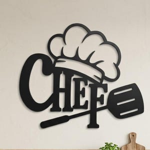 Op de afbeelding: Een zwart metalen wanddecoratie met het woord CHEF in dikke hoofdletters, bekroond met een koksmutspictogram en een spatelafbeelding die in het ontwerp is geïntegreerd. Het bord is aan een muur boven een houten plank in een keuken bevestigd.