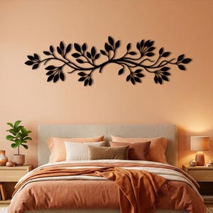 May include: A black metal wall art piece featuring a horizontal, sprawling branch design with numerous small, oval-shaped leaves. The decorative sculpture is mounted centrally on a solid, light peach-coloured wall above a bed headboard.