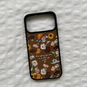 May include: A brown phone case with a floral pattern featuring sunflowers, roses, and other flowers. The case has a black border and the text "Everything you lose is a step you take" in white.